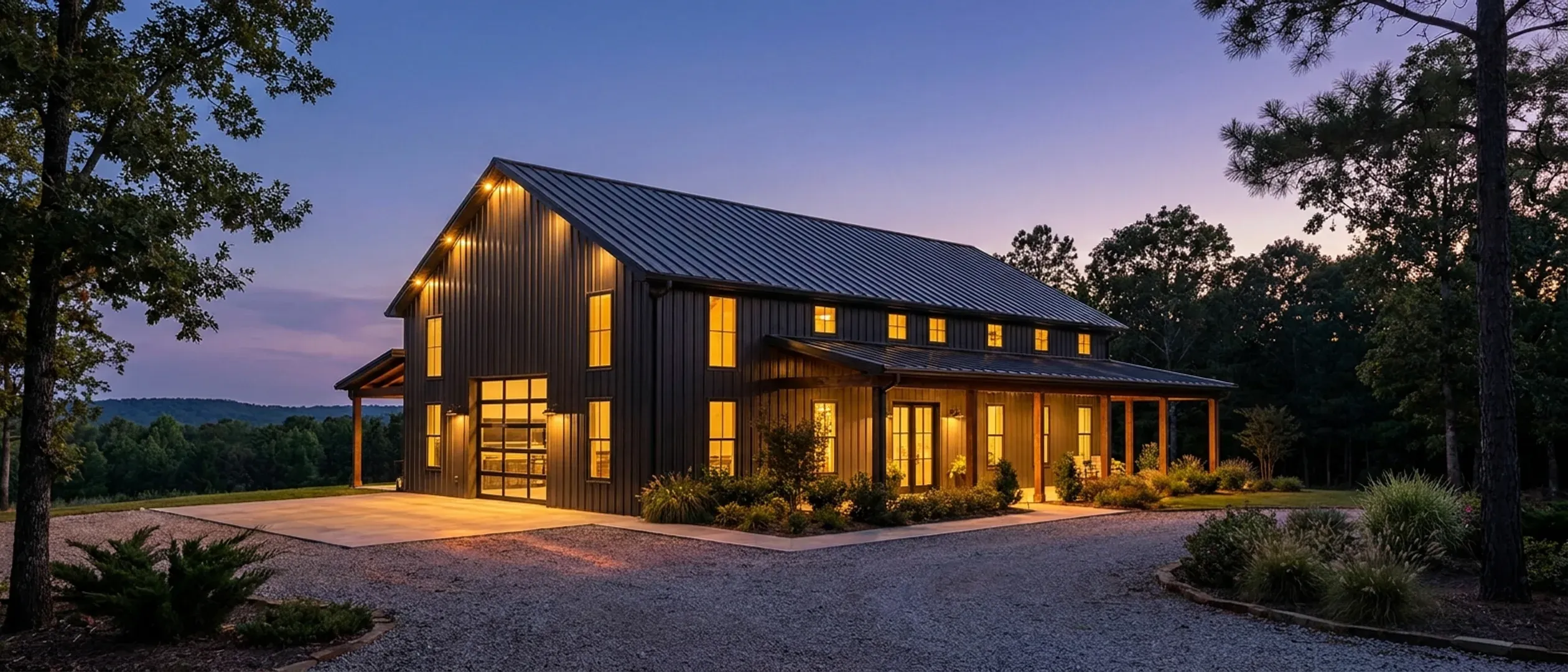 A barndominium at dusk with warm light glowing from every window and exterior lighting along the porch and roofline against a deep blue twilight sky.