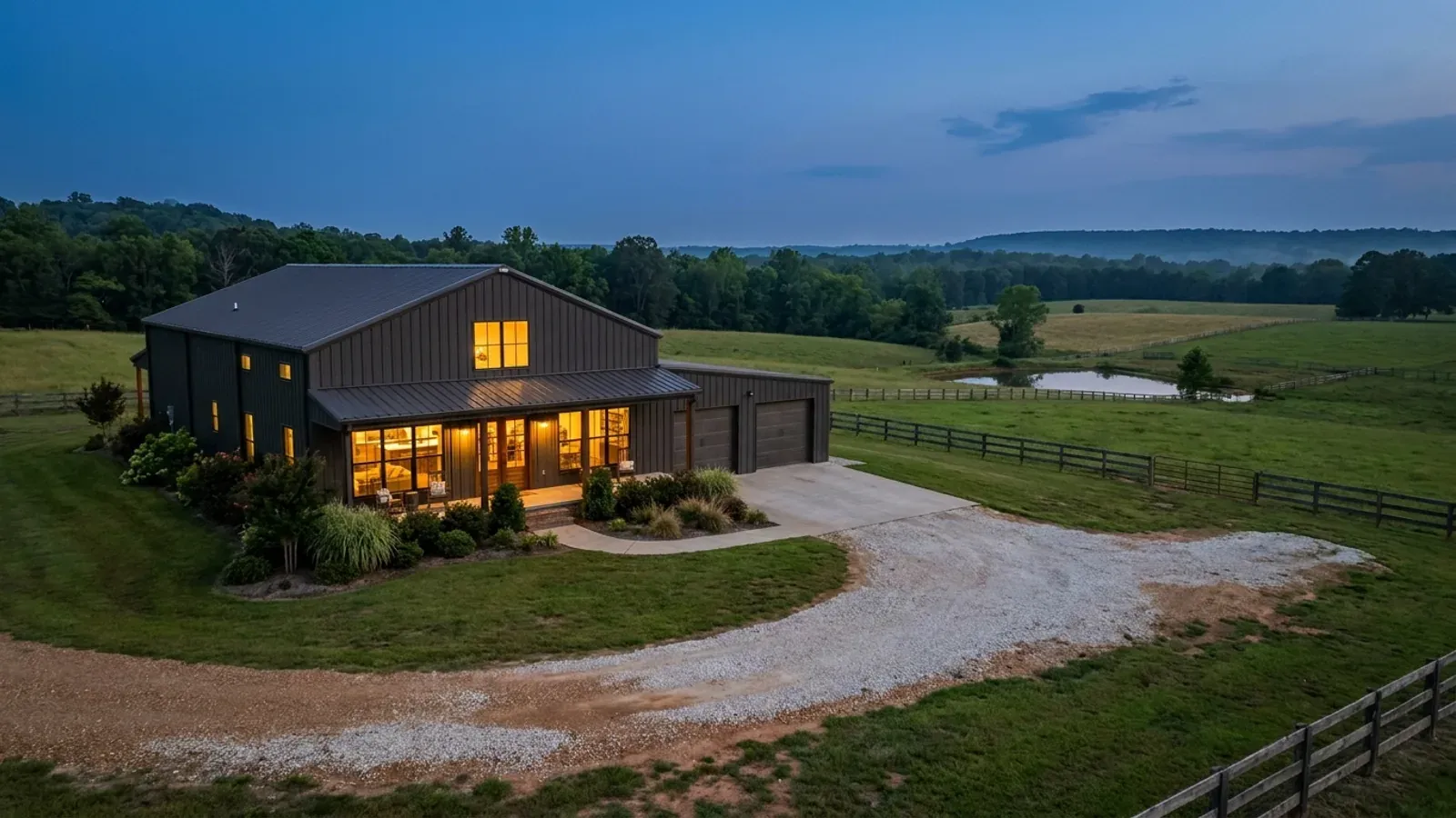 A finished barndominium at dusk with warm interior lighting glowing through its large windows.