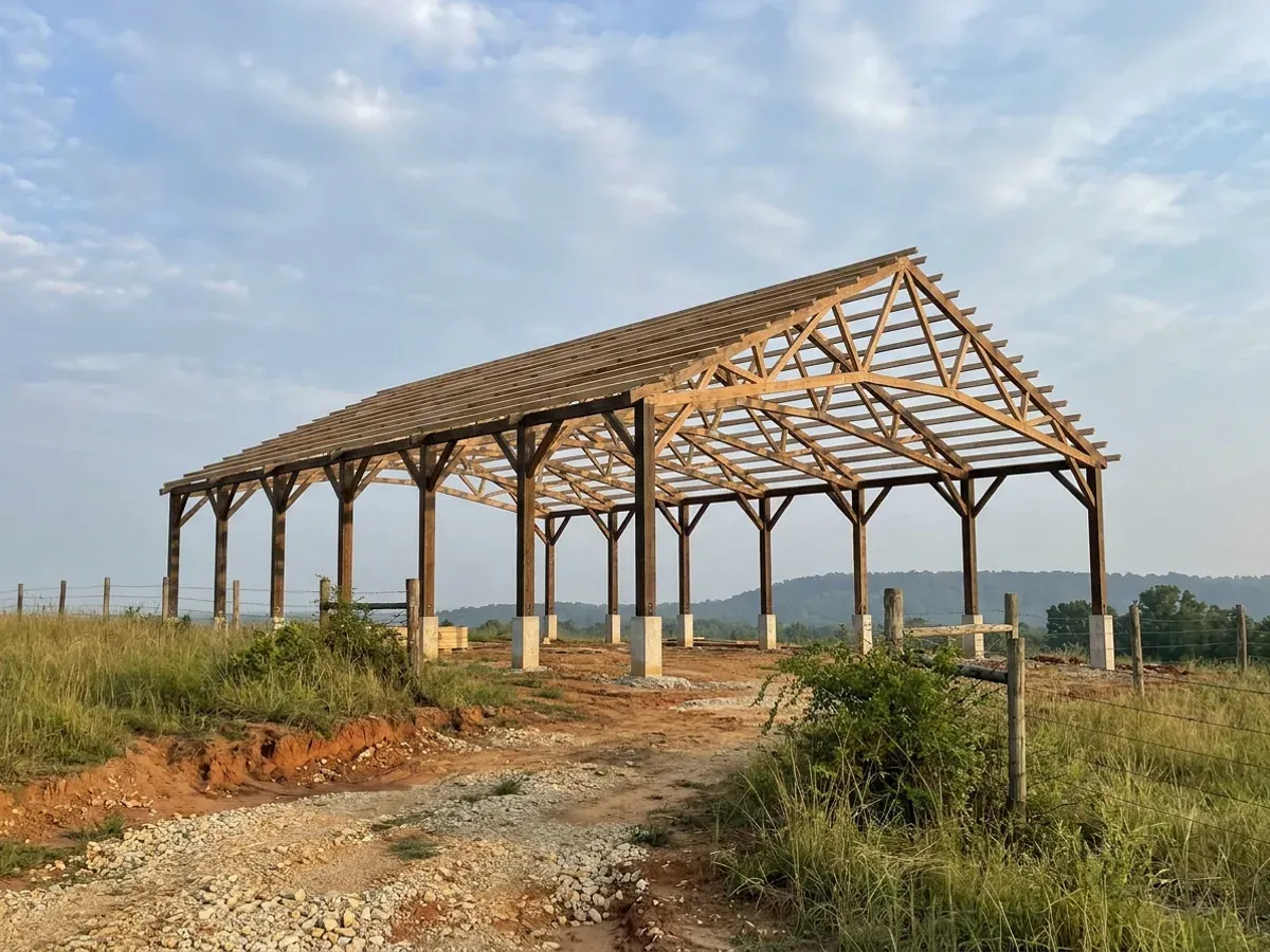 A post-frame home under construction, showing laminated timber columns on concrete piers and wide-span roof trusses with no interior bearing walls.