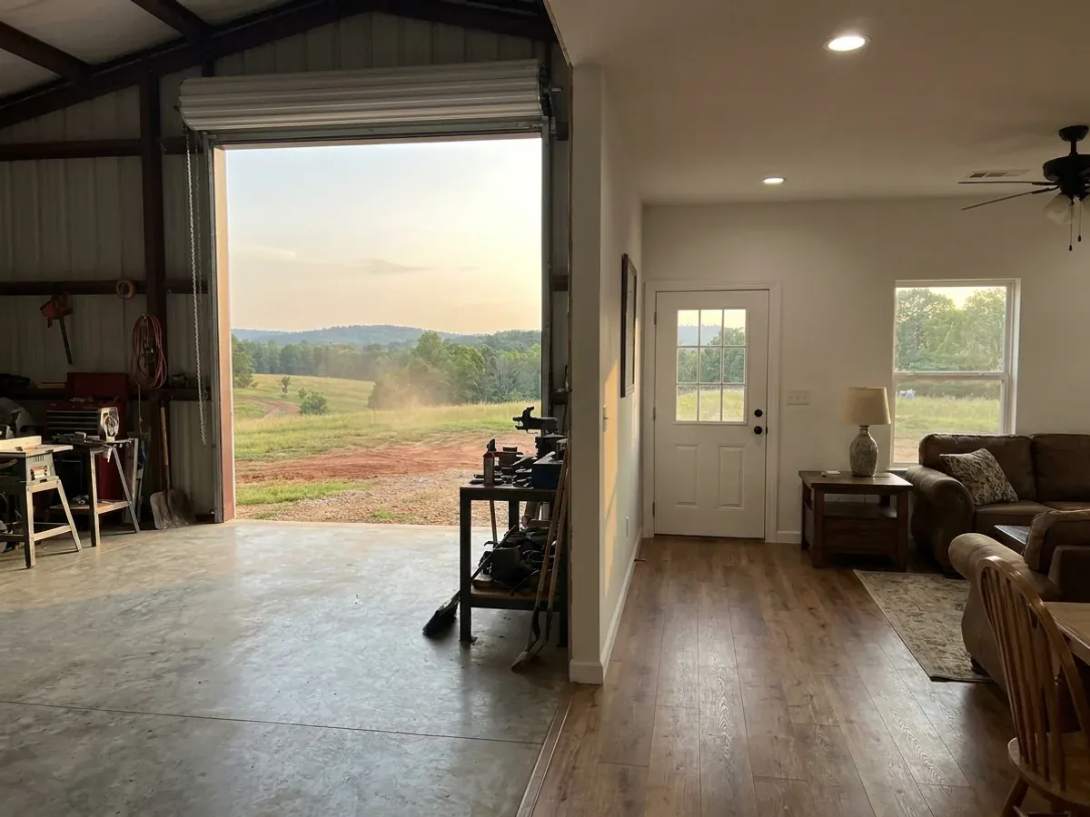 The interior of a shouse where a finished living area with warm flooring meets a shop bay with polished concrete and a tall roll-up door.